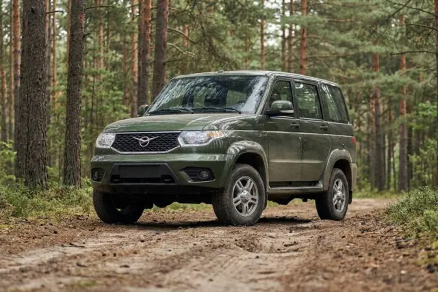 A dark green five-door SUV is parked on a dirt track surrounded by a dense forest of tall pine trees. It has a winged